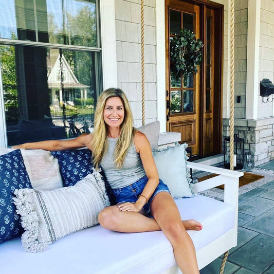 Woman sitting on a white porch swing bed with decorative pillows in front of a house.