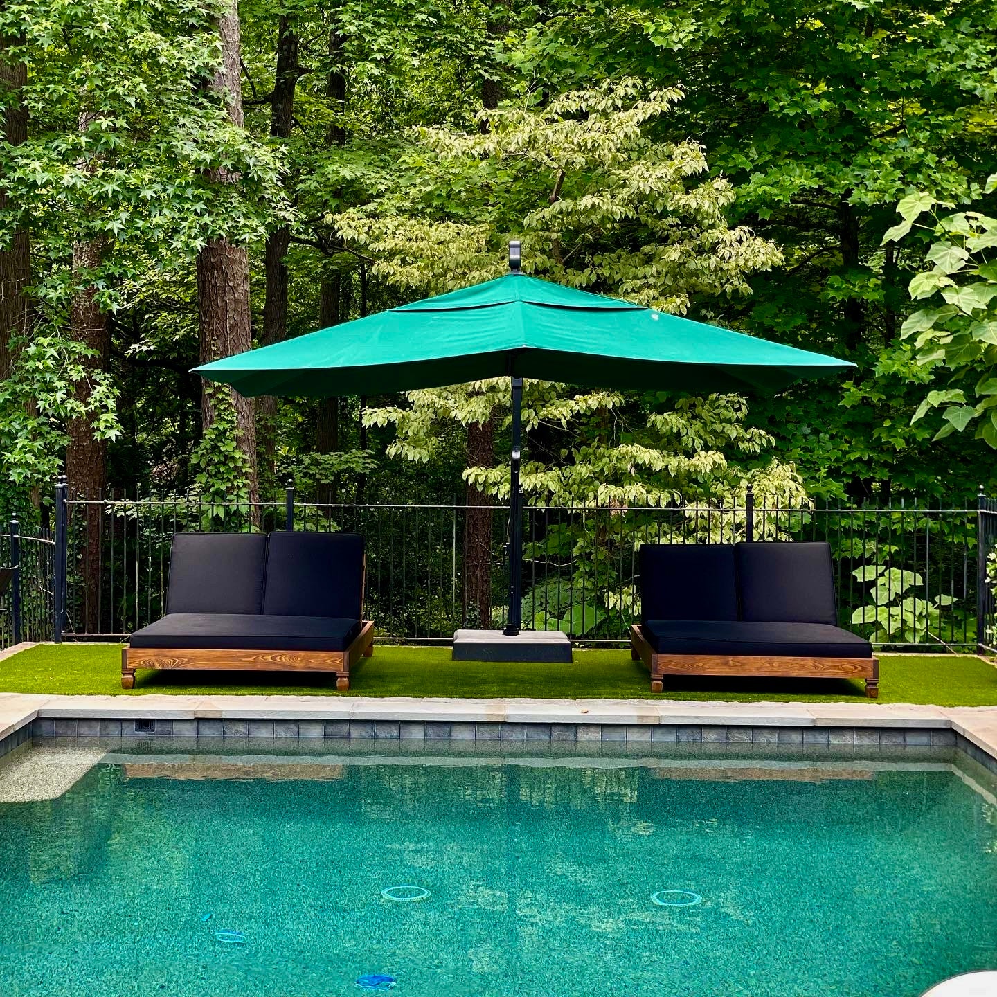 Two lounge chairs with black cushions under a green umbrella by a pool with a forest background
