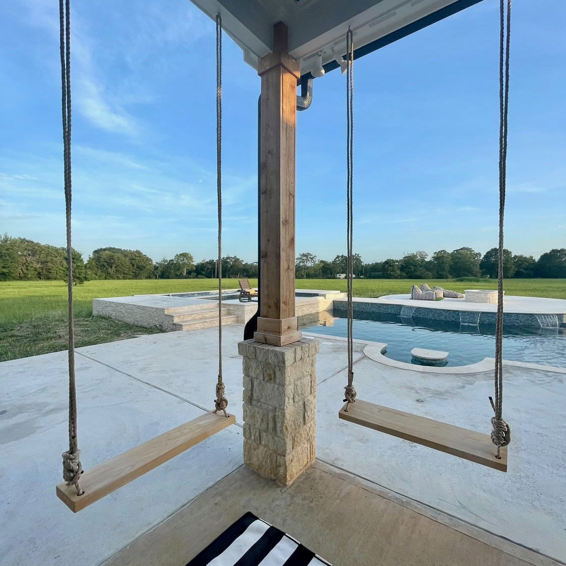 Wooden swings hanging from a porch with a pool and garden view.